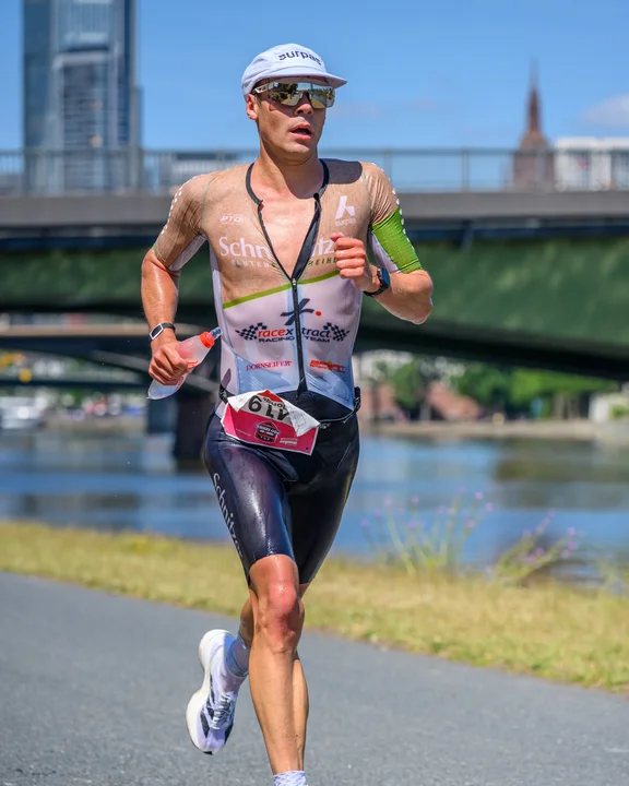 Der Triathlet Jonas Hoffmann läuft mit der Startnummer 6 auf einem Asphaltweg entlang eines Flusses. Er trägt einen gesponserten Trisuit mit den Logos „Schnitzer" und „racex Racing Team", dunkle Laufshorts sowie weiße Socken und Laufschuhe. In der Hand hält er eine Trinkflasche, das Gesicht ist durch Sonnenbrille und weiße Laufmütze geschützt. Im Hintergrund sind eine Brücke, ein Hochhaus und ein Kirchturm bei strahlendem Sonnenschein zu erkennen.