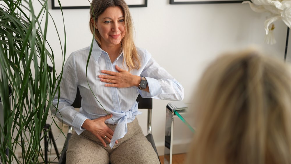 Eine l&auml;chelnde Therapeutin sitzt einer Patientin gegen&uuml;ber und demonstriert eine Atem&uuml;bung: eine Hand liegt auf der Brust, die andere auf dem Bauch. Der Raum ist hell und freundlich eingerichtet.
