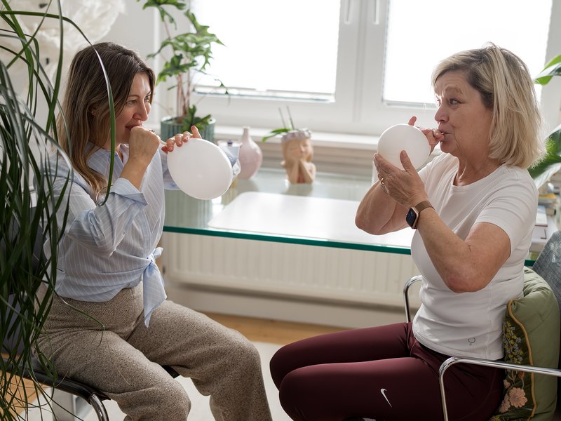 Therapeutin und Patientin &uuml;ben gemeinsam eine Atem&uuml;bung mit wei&szlig;en Luftballons in einem freundlichen Therapieraum.