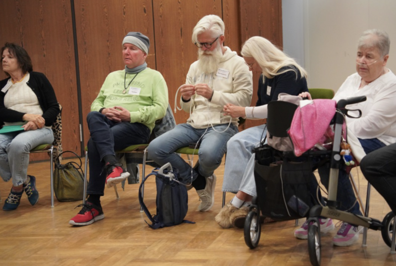 Eine Veranstaltung des Lungenfibrose e.V. F&uuml;nf Personen sitzen in einem Veranstaltungsraum. Eine Teilnehmerin mit Sauerstoffger&auml;t und Rollator ist dabei. Zwei Teilnehmende machen sich mit einem mobilen Sauerstoffger&auml;t mit Schlauch vertraut. Das Ger&auml;t befindet sich in einem kleinen Rucksack.