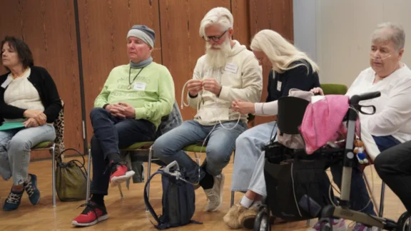 Eine Veranstaltung des Lungenfibrose e.V. F&uuml;nf Personen sitzen in einem Veranstaltungsraum. Eine Teilnehmerin mit Sauerstoffger&auml;t und Rollator ist dabei. Zwei Teilnehmende machen sich mit einem mobilen Sauerstoffger&auml;t mit Schlauch vertraut. Das Ger&auml;t befindet sich in einem kleinen Rucksack.